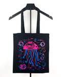 A black canvas tote bag hanging on a black hanger against a white background, featuring a vibrant neon-style graphic of a glitched pink and blue jellyfish.