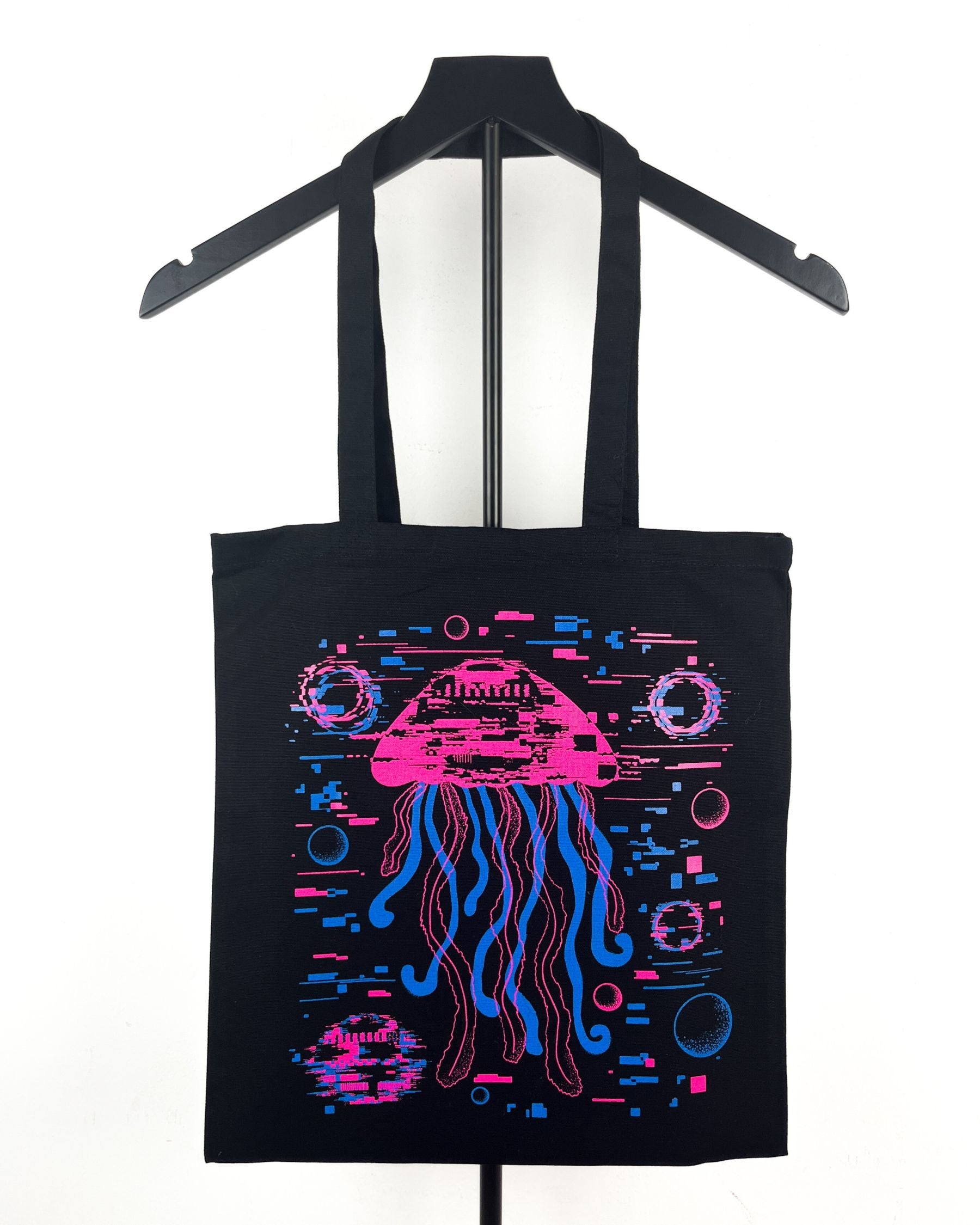 A black canvas tote bag hanging on a black hanger against a white background, featuring a vibrant neon-style graphic of a glitched pink and blue jellyfish.