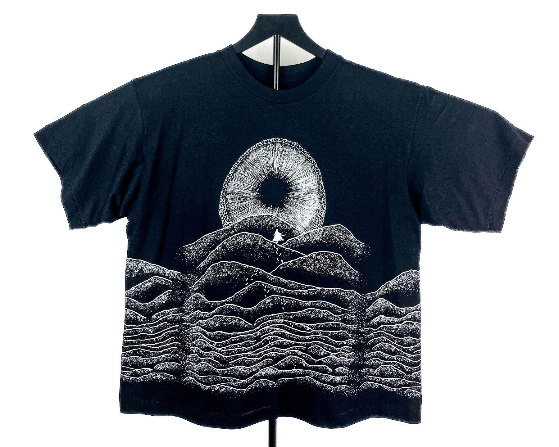 A black short-sleeve t-shirt displayed on a hanger against a white background, featuring a white graphic print of a Shai-hulud mouth from Dune and layered soundwave-style hills.