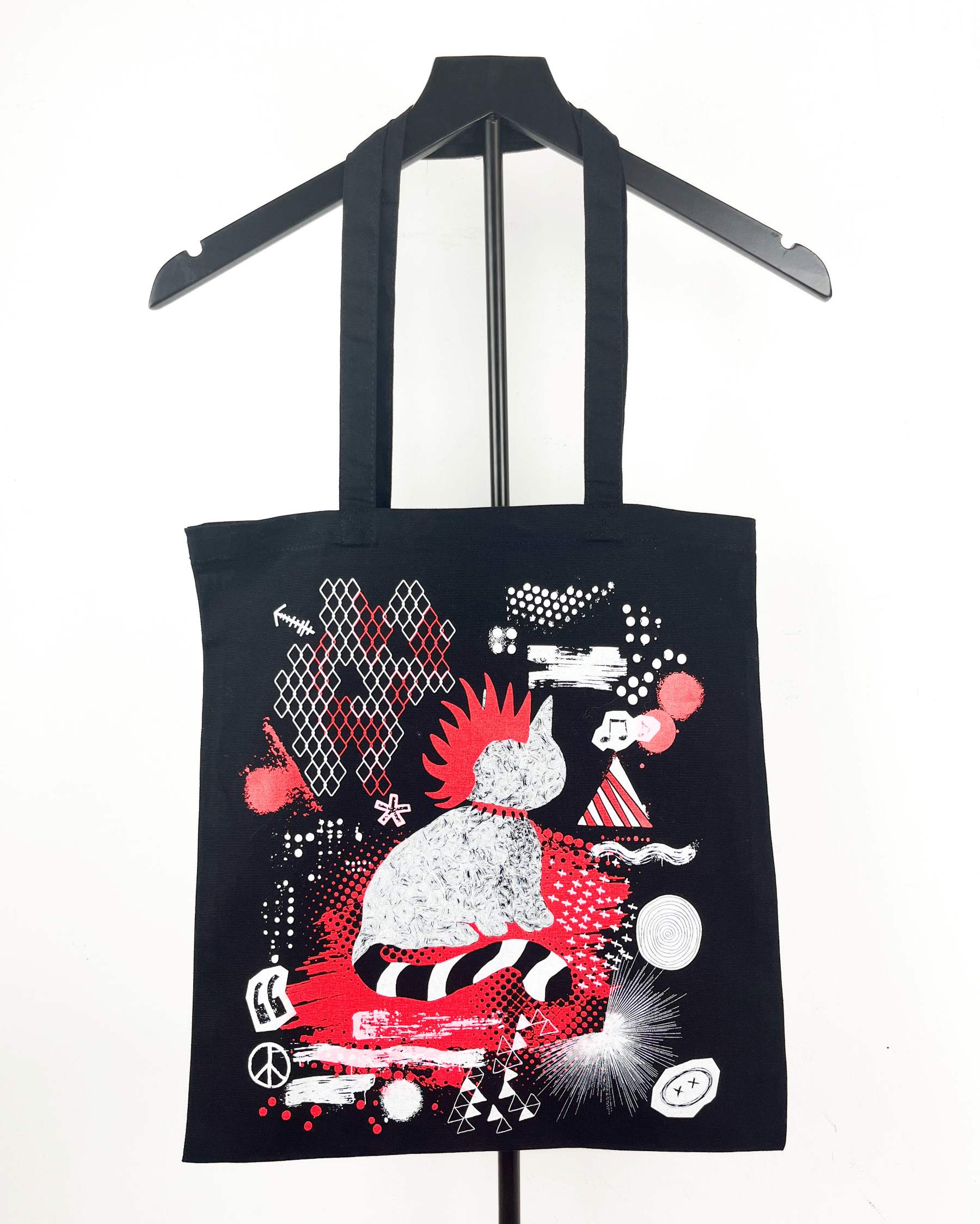 A black canvas tote bag hanging on a black hanger against a white background, featuring a colorful graphic print of a stylized clown in red, white, and black.