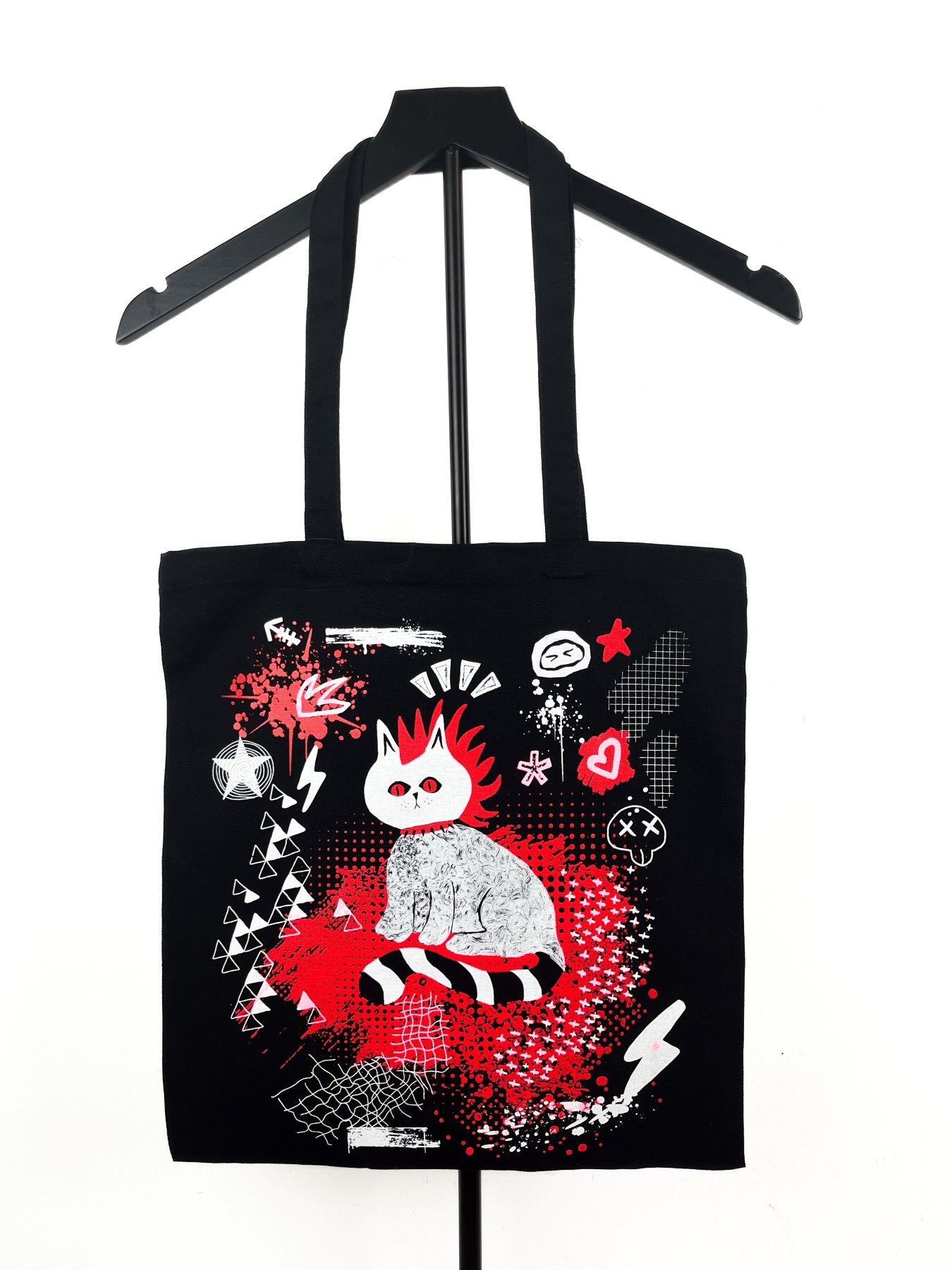 A black canvas tote bag with a graphic print of a stylized white cat with red hair, displayed on a black hanger against a white background.