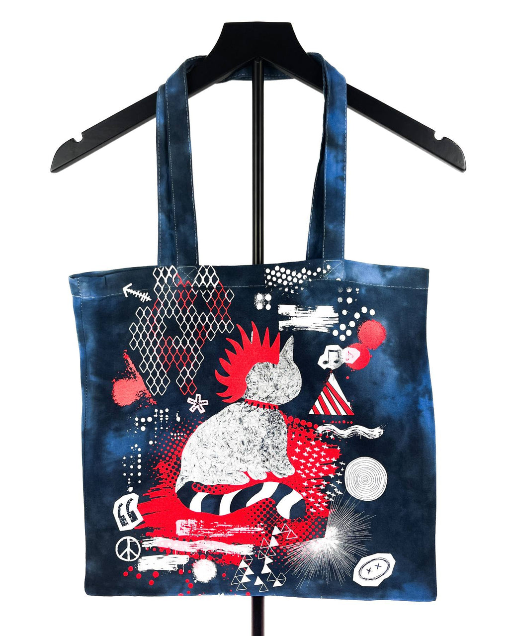 A dark blue tote bag with a black handle, featuring a vibrant graphic print of a cat in a red and white striped outfit surrounded by red, white, and black abstract patterns, displayed on a black hanger against a white background.