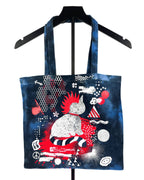 A dark blue tote bag with a black handle, featuring a vibrant graphic print of a cat in a red and white striped outfit surrounded by red, white, and black abstract patterns, displayed on a black hanger against a white background.