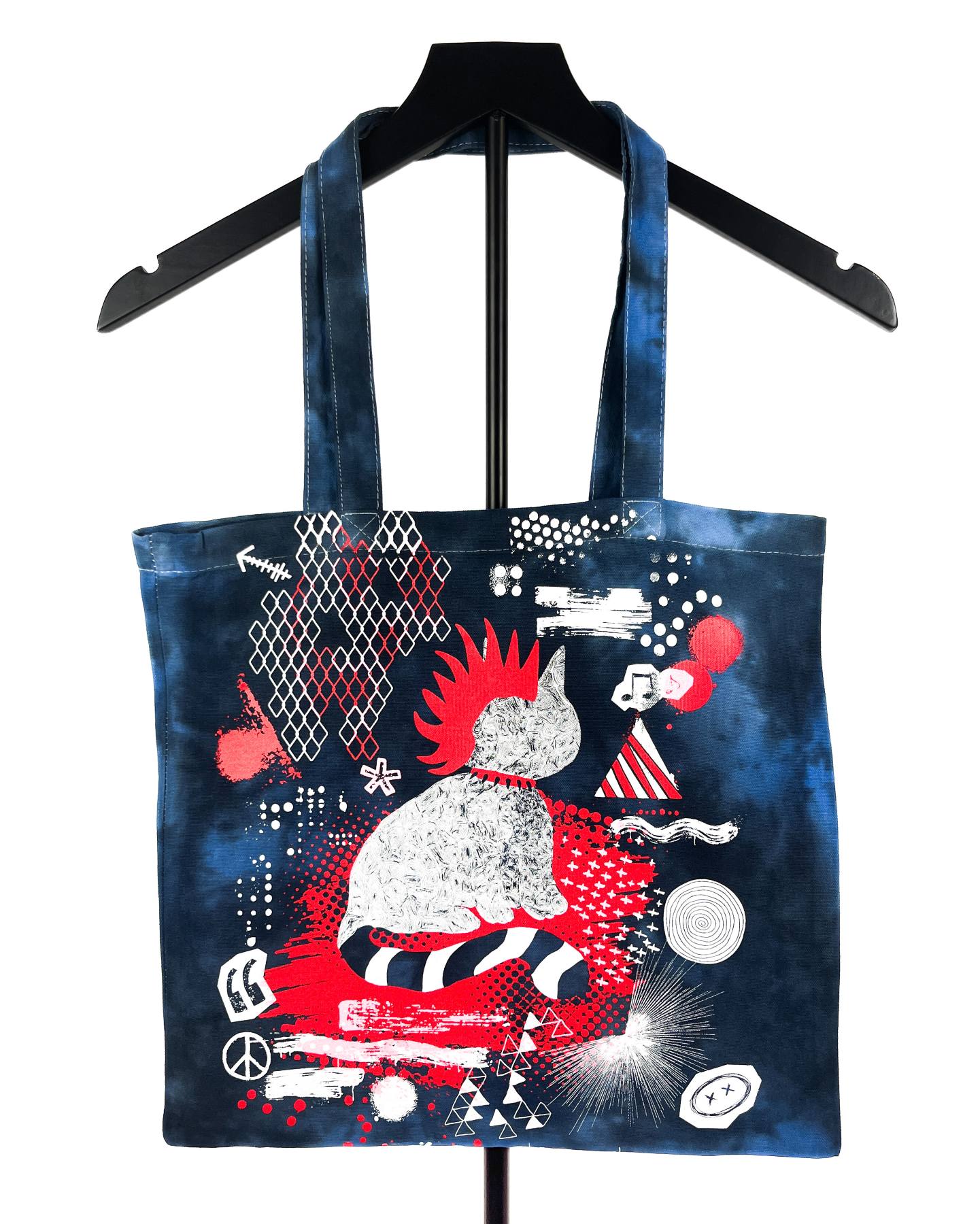 A dark blue tote bag with a black handle, featuring a vibrant graphic print of a cat in a red and white striped outfit surrounded by red, white, and black abstract patterns, displayed on a black hanger against a white background.