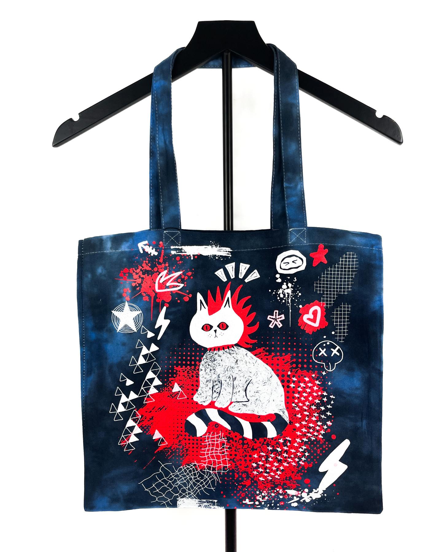 A dark blue tie-dye canvas tote bag with a graphic print of a white cat with red hair, displayed on a black hanger against a white background.