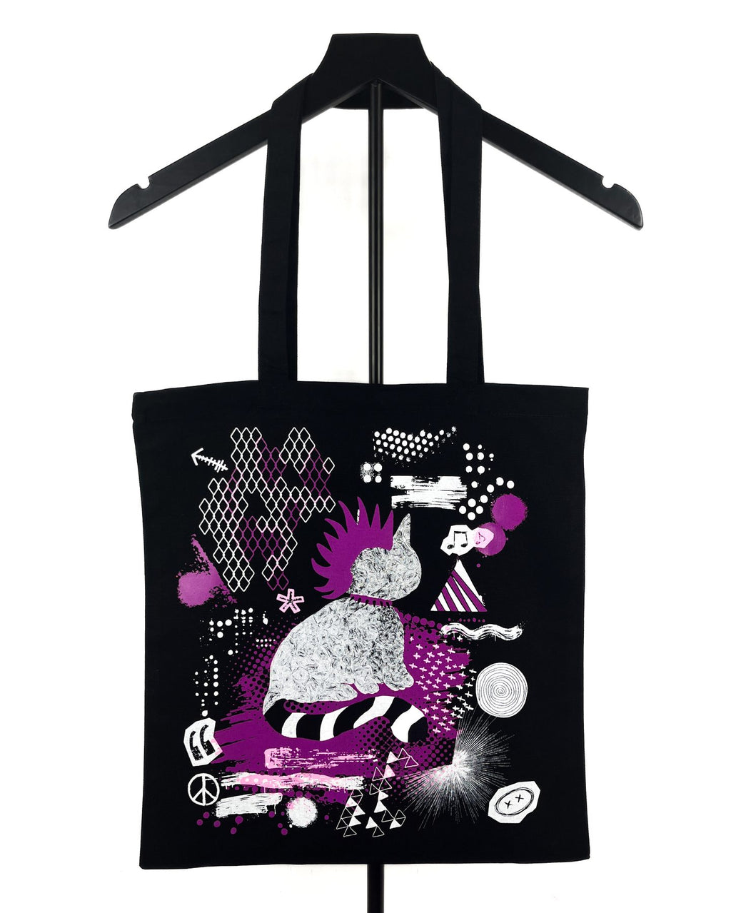A black canvas tote bag with long black shoulder straps, hanging on a black hanger against a white background. The bag features a colorful graphic print in purple, white, and black, depicting a stylized figure and abstract shapes.