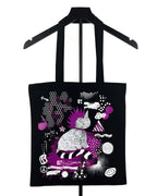 A black canvas tote bag with long black shoulder straps, hanging on a black hanger against a white background. The bag features a colorful graphic print in purple, white, and black, depicting a stylized figure and abstract shapes.