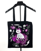 A black canvas tote bag with a gothic-style graphic print of a white cat with purple hair, hanging on a black hanger against a white background.