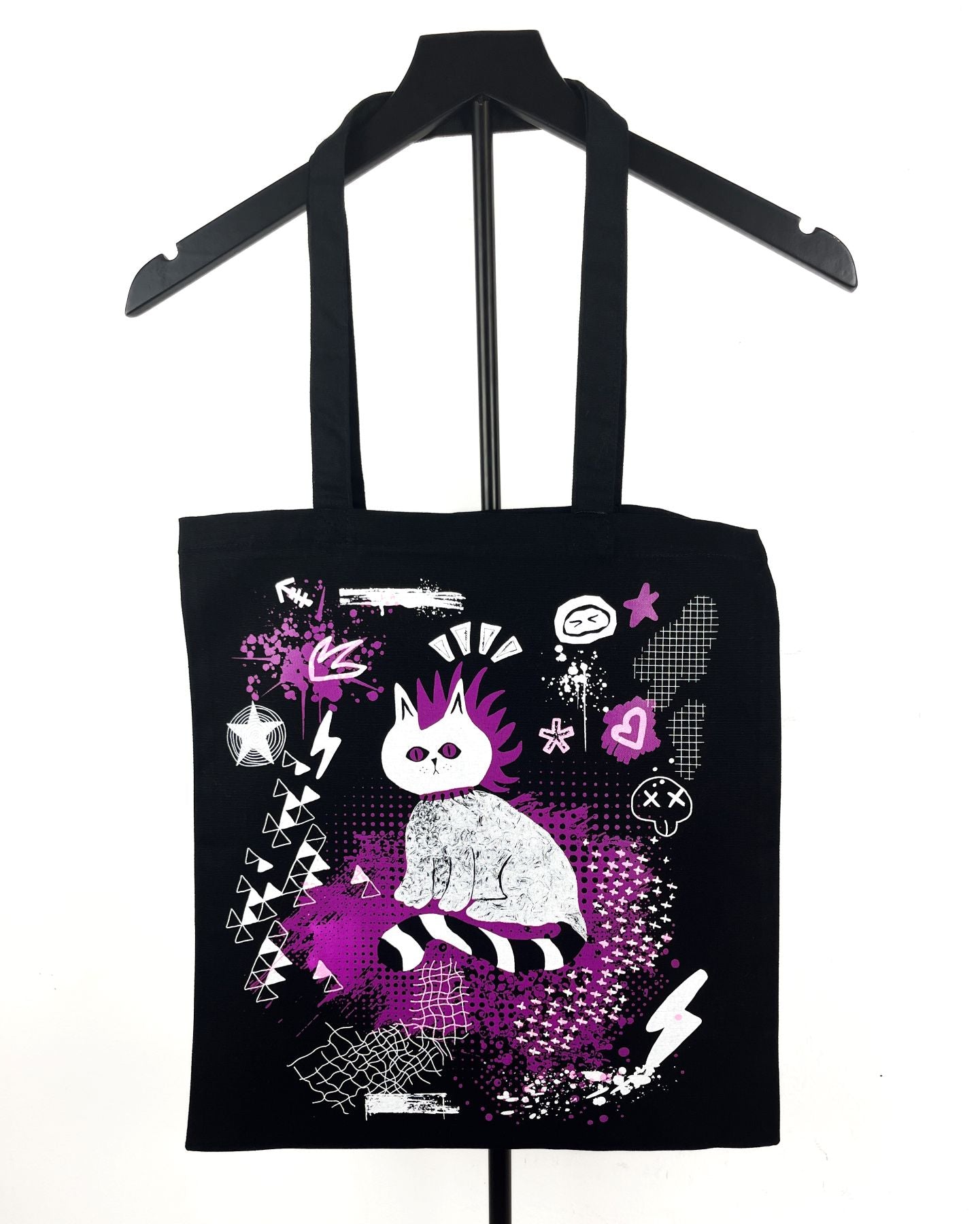A black canvas tote bag with a gothic-style graphic print of a white cat with purple hair, hanging on a black hanger against a white background.