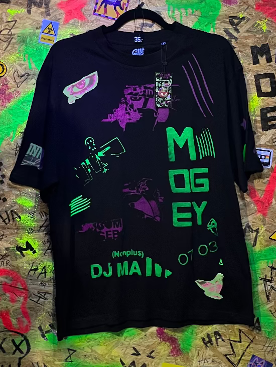 Black t-shirt with colorful graphics and text hanging against a graffiti-covered wall.