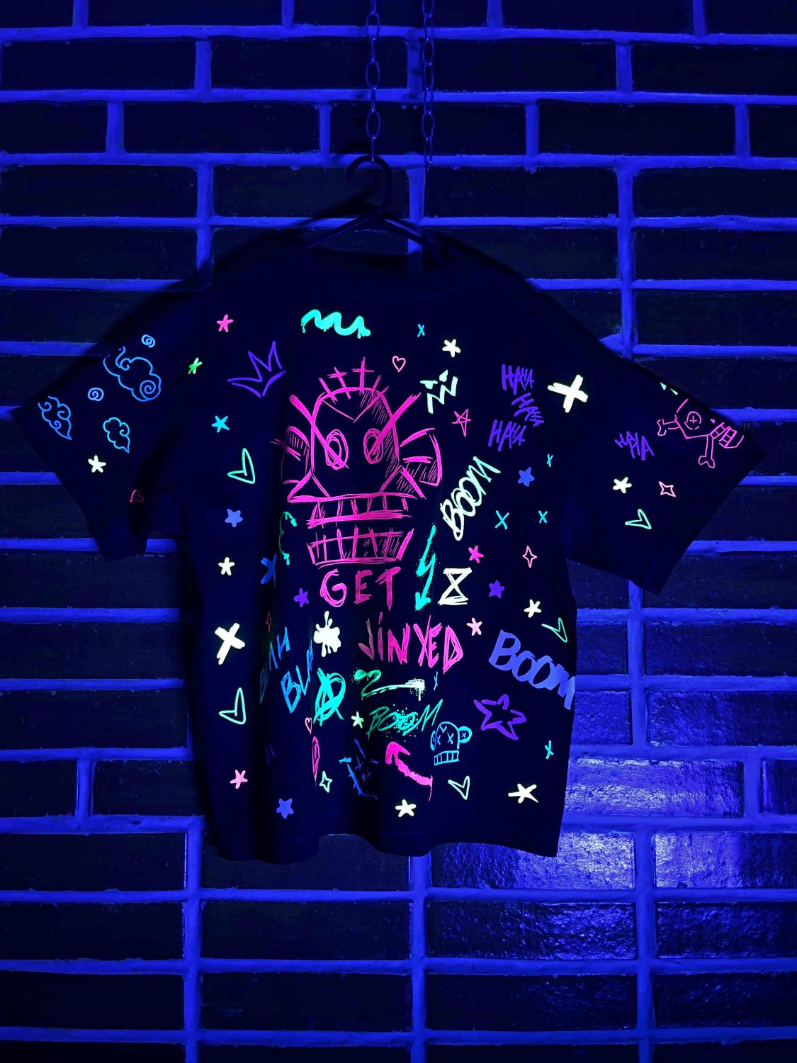 Black t-shirt with colorful designs glowing under black light against a brick wall.