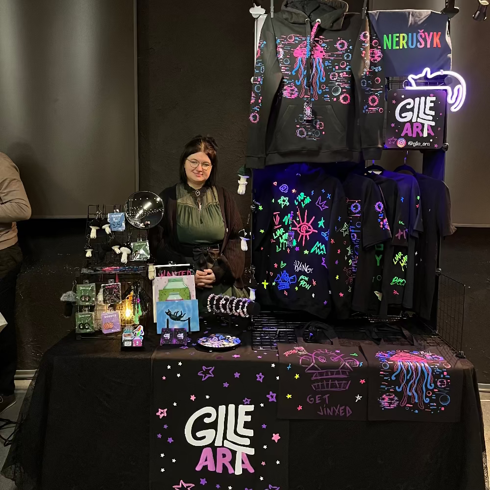Person standing behind a table with merchandise displaying 'Gile Art' at an event.