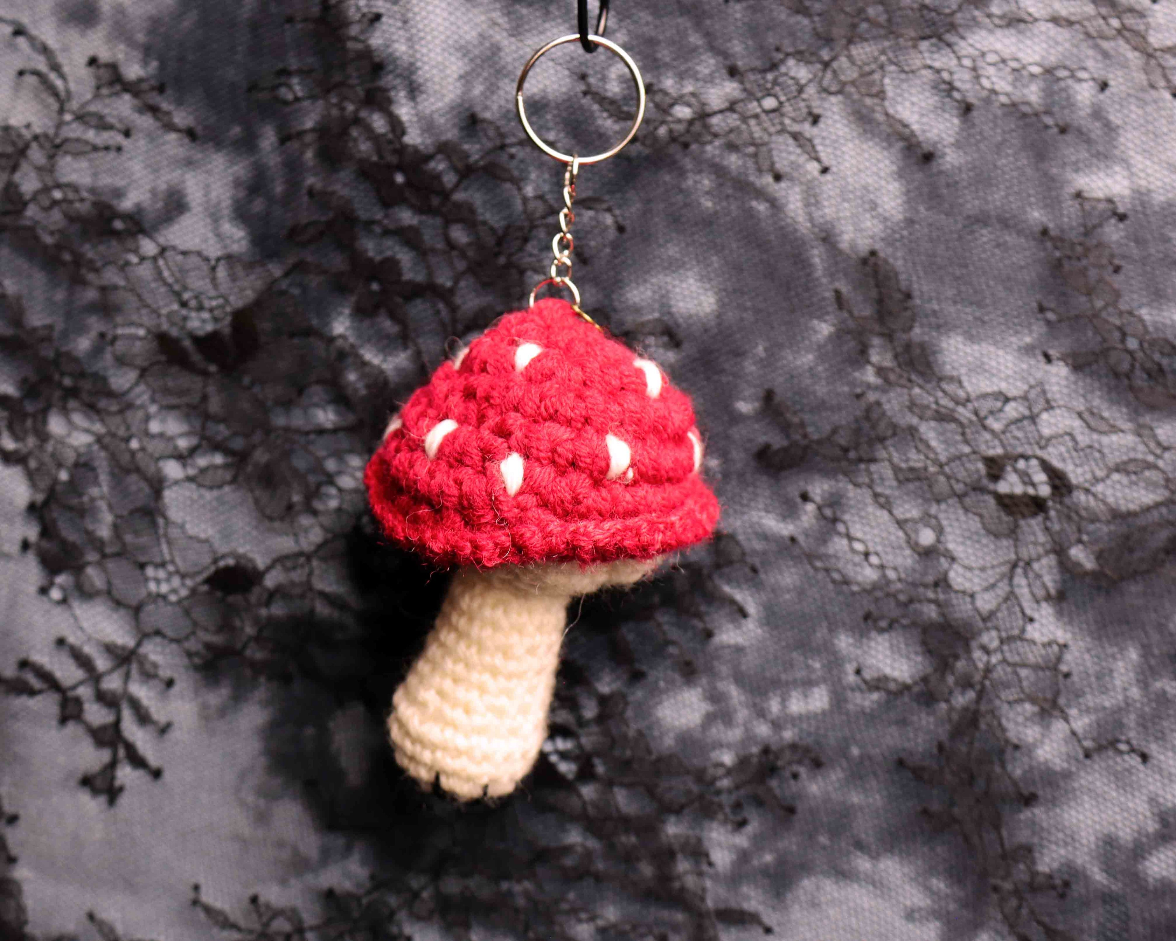 Crocheted mushroom keychain with red cap and beige stem on a textured gray background