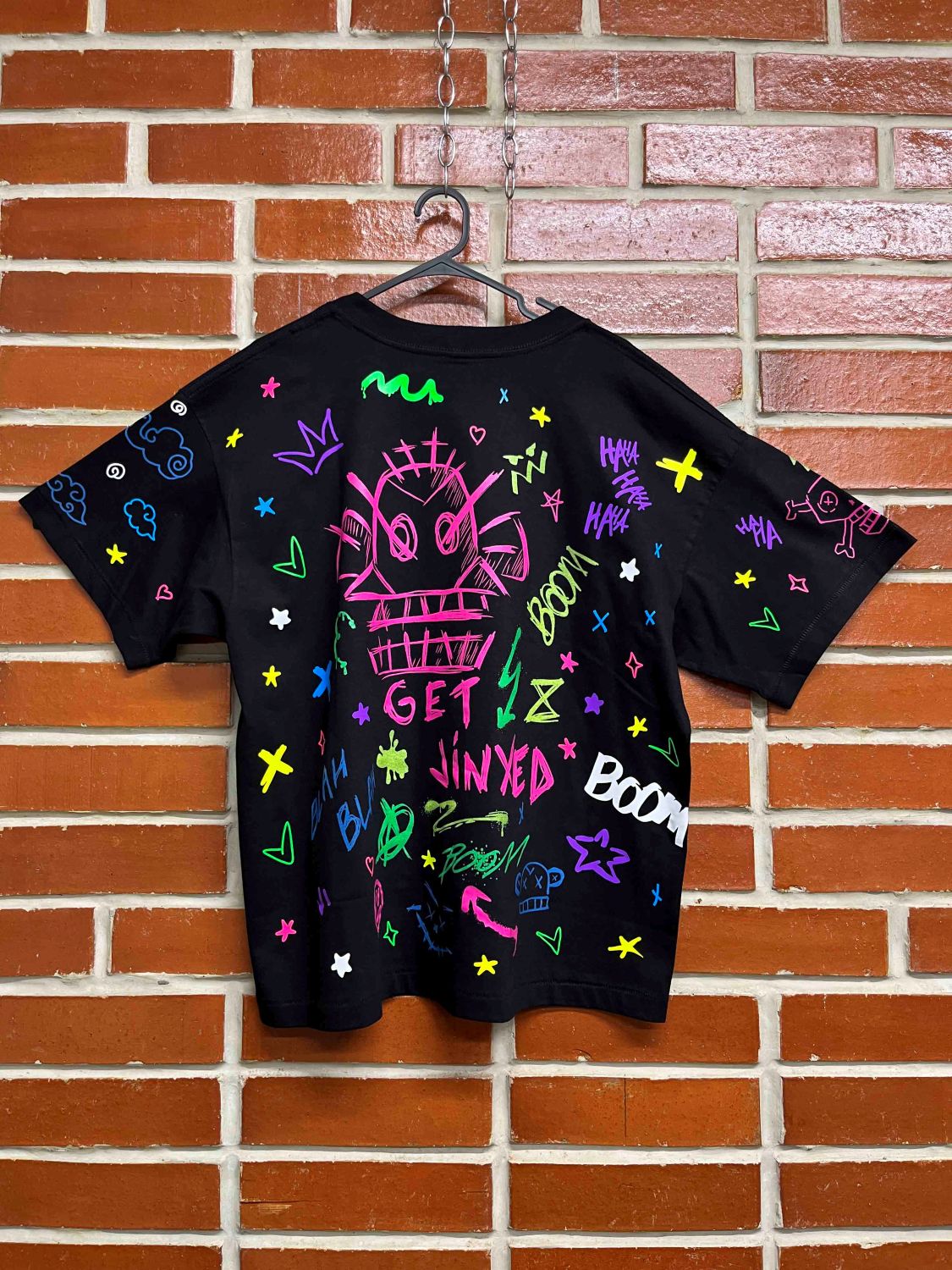 Black t-shirt with colorful graphics and text on a hanger against a brick wall.